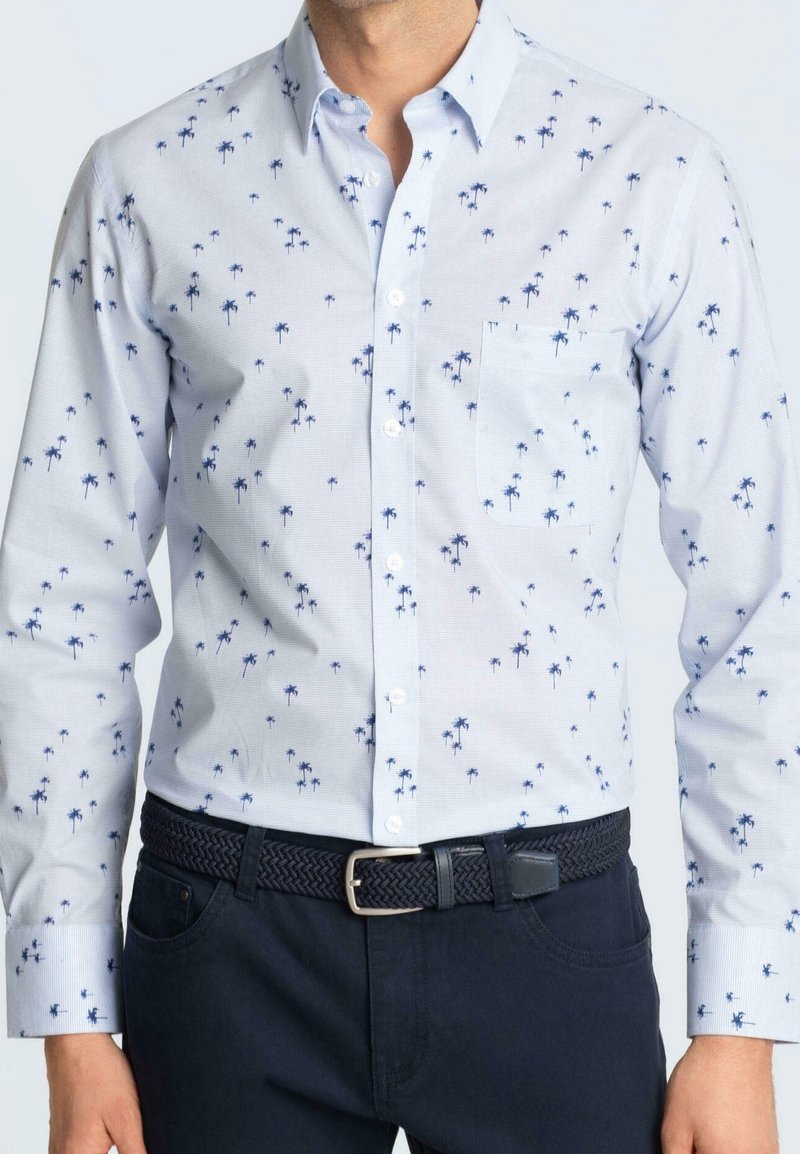 Homme portant une chemise bleu clair à boutons avec un imprimé de palmiers bleu foncé, ceinture tissée sombre et pantalon foncé, debout les mains le long du corps.