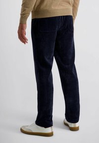 Man in een marineblauwe corduroy broek, beige trui en witte sneakers met gumzolen, staand tegen een effen lichte achtergrond.