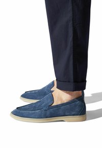 Blaue Wildlederschlüpfschuhe mit einer abgerundeten Zehe, minimalen Nähdetails und einer hellen Gummisohle. Getragen mit navyfarbenen Hosen.