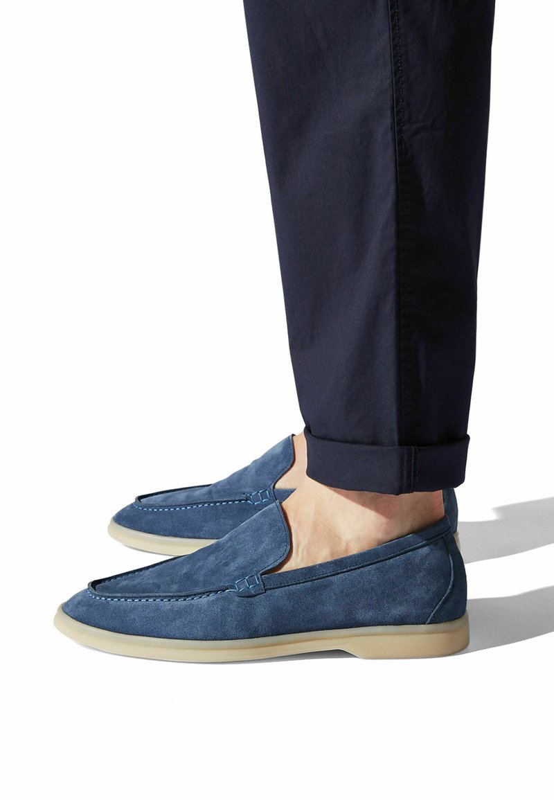 Blaue Wildlederschlüpfschuhe mit einer abgerundeten Zehe, minimalen Nähdetails und einer hellen Gummisohle. Getragen mit navyfarbenen Hosen.