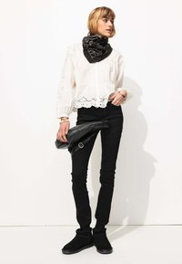 Blouse blanche avec bord en dentelle, jean skinny noir, foulard noir à motifs, et bottines noires, tenant une veste en cuir noir.