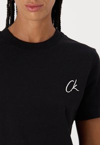 Černé bavlněné tričko s kulatým výstřihem, krátkými rukávy a bílým vyšívaným logem "Ck" na levé straně hrudi. Hladká textura, klasický střih.
