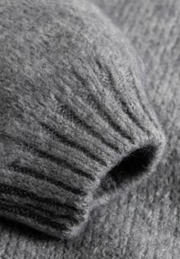 Pilkas megztinis iš vilnos artimas vaizdas, su ribuotais rankogaliais ir minkšta faktūra su švelniu blizgesiu, išryškinantis audinio šilumą ir detales.