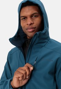 Turkusowa kurtka softshell z zamkiem błyskawicznym w kieszeni, wysokim kołnierzem i regulowanym kapturem. Posiada przeszycia i gładką teksturę.