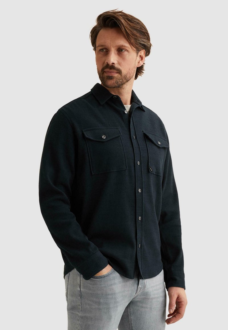 Homme portant une chemise bleu marine foncé boutonnée avec des poches poitrine et un jean gris clair, debout avec une main dans la poche, regardant sur le côté.