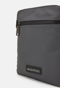 Borsa in nylon grigia con una texture liscia, dotata di una tasca con zip e un'etichetta in pelle nera per il brand.