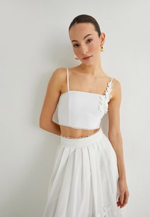 Top corto blanco con tirantes delgados, con apliques florales en el lado izquierdo y dobladillo ondulado, combinado con una falda blanca de plisados.