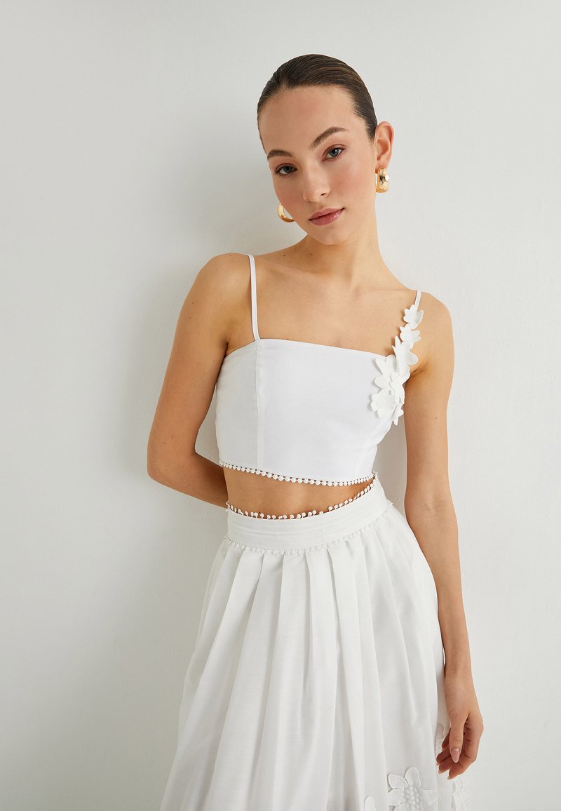 Witte crop top met dunne banden, bloemenapplicatie aan de linkerkant en gegolfde zoom, gecombineerd met een witte plissé rok.