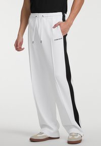 Weiße Jogginghose mit schwarzem Seitenstreifen, ausgestattet mit einem elastischen Taillenbund und Kordelzug. Sie verfügt über eine Seiten Tasche und das Logo 'GUESS JEANS'.