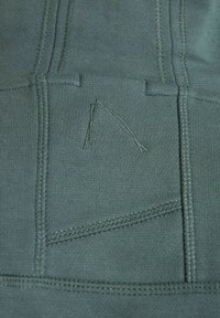 Tessuto verde scuro con cuciture testurizzate, un sottile dettaglio di ricamo triangolare e un design che ricorda una tasca, visto da vicino.