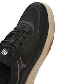 Zapatilla deportiva negra con parte superior de malla, sobrecapas sintéticas y suela de goma beis. Presenta cordones negros y un discreto detalle de raya dorada.
