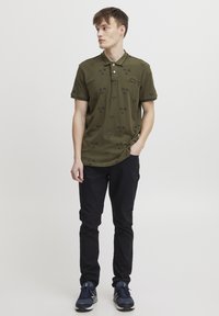 Oliven grønn polo skjorte med svart palme mønster, korte ermer, og tre-knappers placket. Kombinert med tettsittende svarte bukser og sneakers.