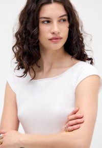 Chemise blanche à manches courtes en tissu doux et lisse avec un col rond. Le modèle pose les bras croisés, mettant en valeur des bagues sur les mains.