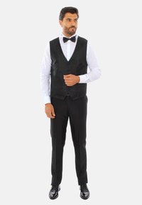 Gilet nero con motivo sottile sopra una camicia bianca, abbinato a pantaloni neri e scarpe eleganti nere lucide; completato con un papillon nero.