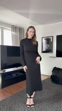 Crna haljina s dugim rukavima i krojem koji prati liniju tijela, izrađena od glatkog tkanja. Uz nju crne sandale s tankim remenčićima oko gležnja.