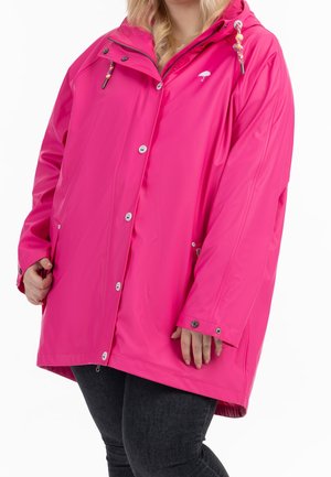 Femme portant une veste imperméable rose vif avec fermeture éclair et boutons, cordons perlés et jean noir.