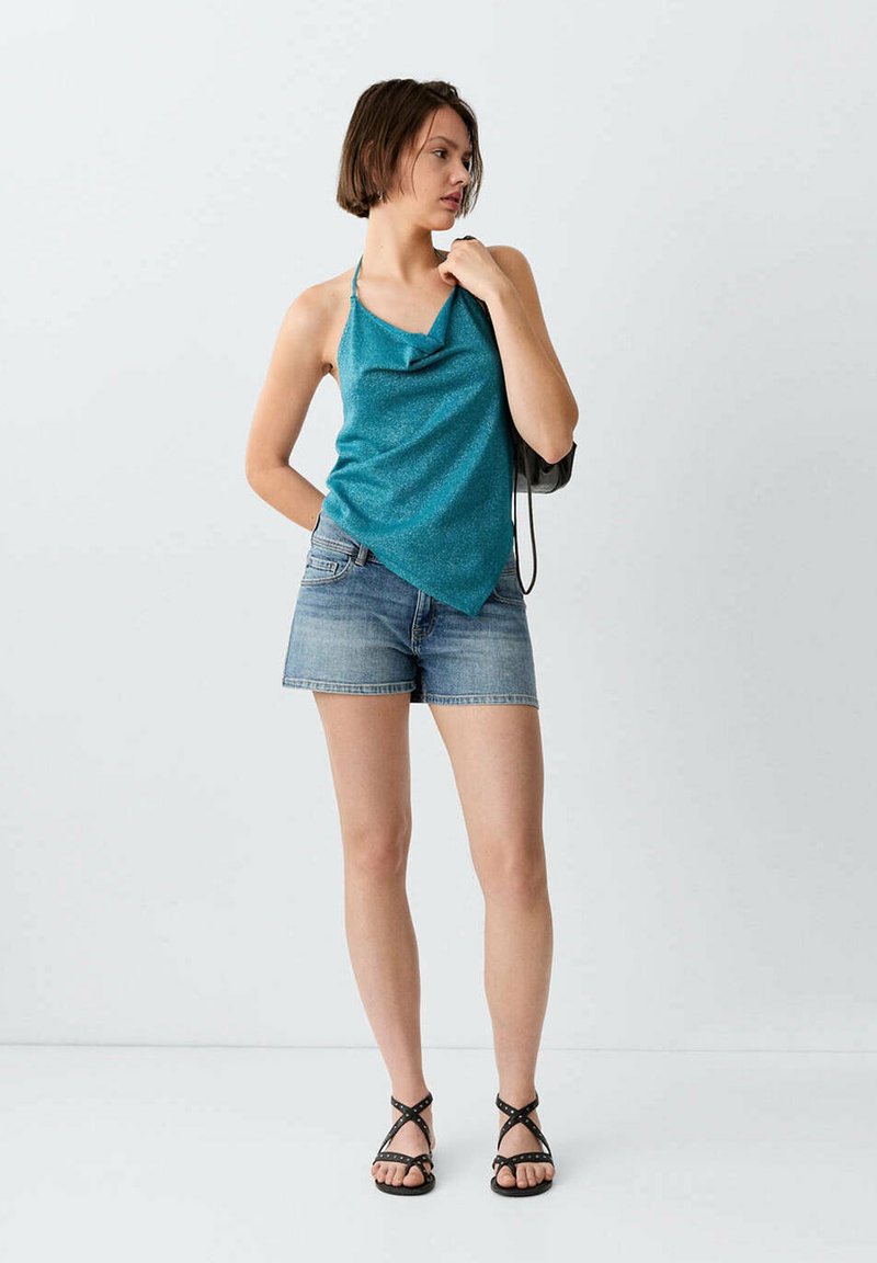 Jeune femme portant un haut dos nu turquoise, un short en jean, des sandales noires à lanières, tenant un petit sac noir, debout devant un fond blanc.