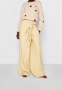 Jasno beżowy sweter z kolorowym haftem owoców, zestawiony z luźnymi, wysokim stanem żółtymi spodniami związanymi paskiem.