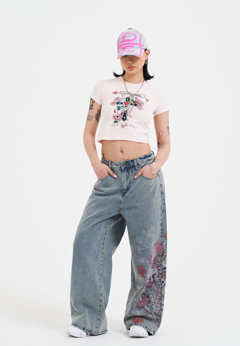 Kort lyserød cropped t-shirt med blomsterprint og tekst, parret med løse blå jeans, der har blomster- og kranietgrafik på det ene ben. Grå kasket.