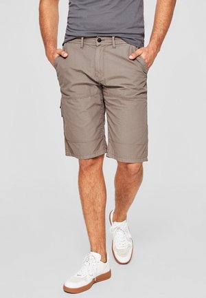 Mann trägt taupefarbene Cargo-Shorts bis zum Knie, graues T-Shirt, weiße Turnschuhe, steht mit den Händen in den Taschen vor einfachem Hintergrund.