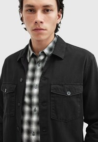 Schwarze Button-Up-Jacke mit zwei Brusttaschen, über einem grau-schwarz karierten Hemd getragen. Weicher Stoff mit einem lässigen Design.