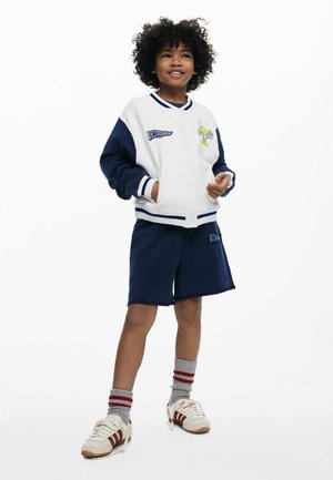 Veste universitaire légère blanche avec manches bleu marine, arborant un graphique et des logos. Associée à un short en éponge bleu marine et des chaussettes rayées.