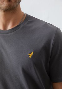 Tricou din bumbac gri, cu guler rotund, maneci scurte și un mic logo brodat cu un păsărele galben în partea stângă a pieptului.