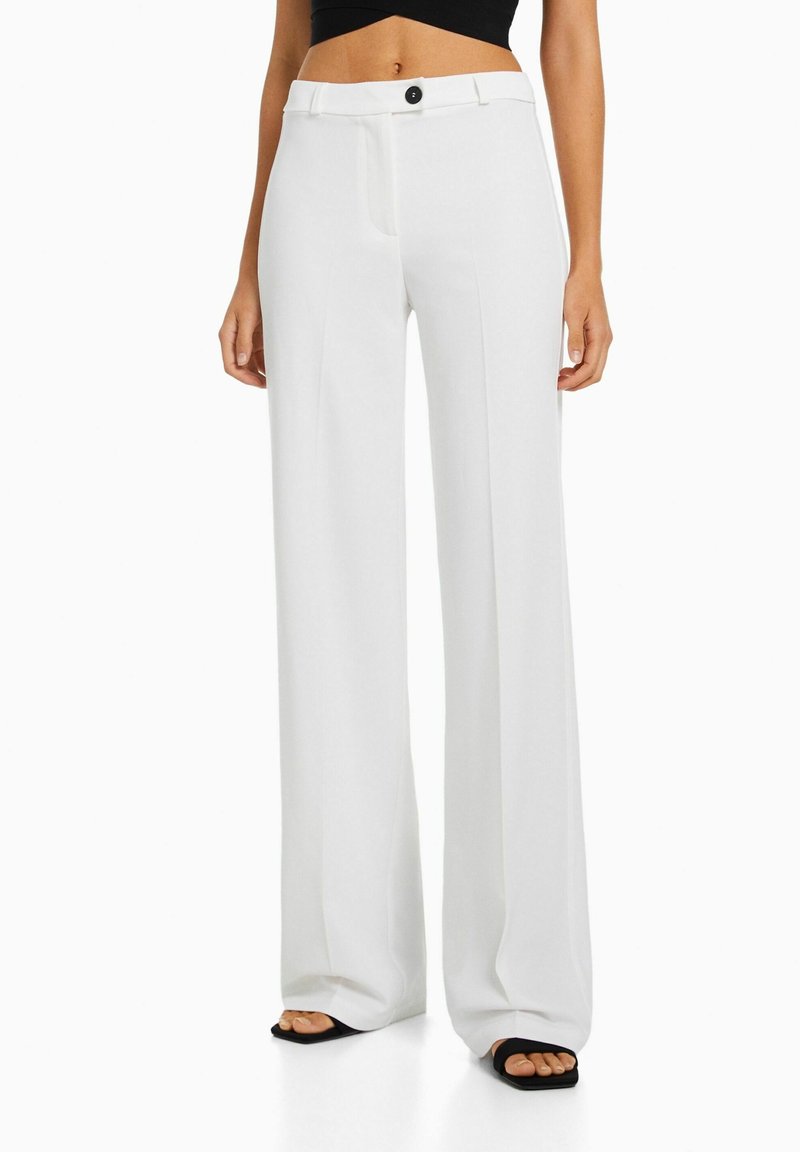 Pantalons larges blancs avec une texture lisse, dotés d'une fermeture à bouton et d'un design ajusté. Portés avec un haut noir ajusté et des sandales.