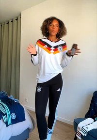 Vrouw die een Duits voetbalshirt en zwarte Adidas legging draagt, gebaart met telefoon in de hand in een slaapkamer met kleding op bed en stoel.