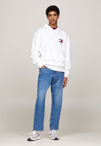 Felpa bianca con tasca frontale, dettagli del logo, abbinata a jeans dritti blu chiaro e sneaker grigie con accenti bianchi.