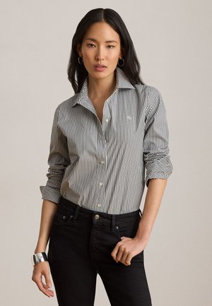 Žena s dugom tamnom kosom koja nosi crno-bijelu prugastu košulju s gumbima, crne traperice, srebrnu narukvicu i naušnice-obruče na neutralnoj pozadini.