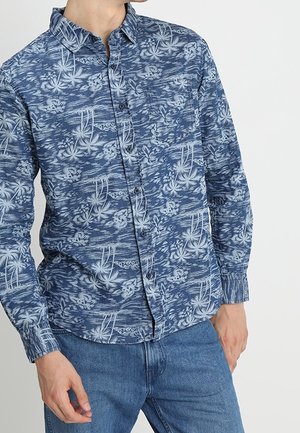 Mand iført en blå langærmet skjorte med hvide tropiske blomsterprint og blå jeans mod en ensfarvet baggrund.