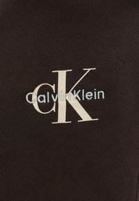 Καφέ υφασμάτινο υλικό με το κείμενο "CalvinKlein" σε γαλάζινη ραφή και χαρακτηριστικό λογότυπο "CK" σε λευκό χρώμα.