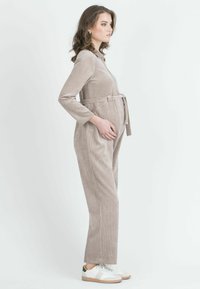 Un mono de pana beige con mangas largas, una cintura ceñida con cinturón y bolsillos laterales, combinado con zapatillas blancas con detalles verdes.