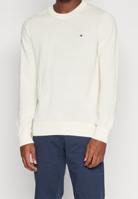 Man in een witte gebreide trui met lange mouwen en een klein logo op de borst, in combinatie met marineblauwe broek, staande tegen een effen achtergrond.