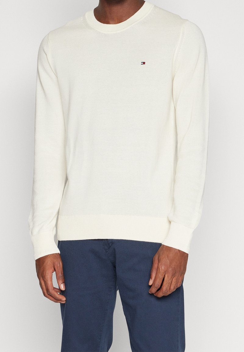 Man in een witte gebreide trui met lange mouwen en een klein logo op de borst, in combinatie met marineblauwe broek, staande tegen een effen achtergrond.