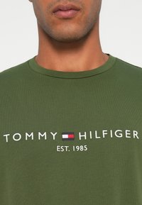 Olivengrøn t-shirt med rund hals, der har hvidt broderet tekst "TOMMY HILFIGER EST. 1985" og et lille logo i rød og blå.