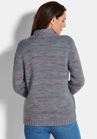 Vrouw die zijwaarts kijkt, gekleed in een multicolore gebreide trui met een sjawl hals en blauwe jeans, tegen een effen lichte achtergrond.