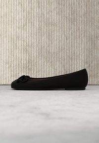 Ballerine noire avec un petit détail en nœud sur le dessus, exposée sur une surface grise sur un fond beige à rayures verticales.