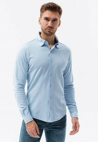 Camisa de manga larga azul claro con cuello abotonado, tejido texturizado y un diseño ajustado, combinada con jeans oscuros.