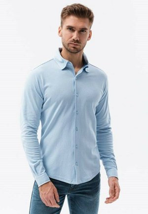 Camisa de manga larga azul claro con cuello abotonado, tejido texturizado y un diseño ajustado, combinada con jeans oscuros.