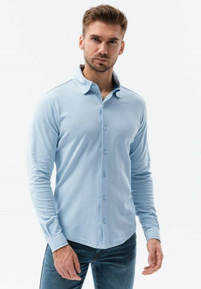 Camisa de manga larga azul claro con cuello abotonado, tejido texturizado y un diseño ajustado, combinada con jeans oscuros.