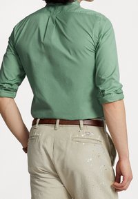 Zielona bawełniana koszula z podwiniętymi rękawami i plisą z tyłu, w zestawie z jasnobeżowymi spodniami z plamami farby i brązowym paskiem.