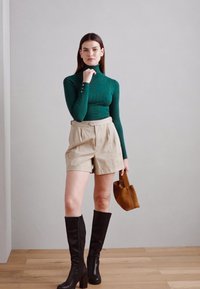 Green ribbed turtleneck, beige high-waisted shorts, knee-high black boots, and a brown handbag. Set against a light gray background.