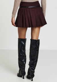 Femme portant une jupe plissée marron avec une fine ceinture noire, un pull en tricot beige et des bottes noires vernies à talons jusqu'aux genoux, se tenant sur un sol blanc.