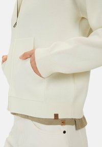 Crème-kleurige gebreide jas met een ritssluiting, geribde manchetten en een zak. Inclusief een logo-tag nabij de zoom, gedragen over beige broeken.