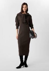 Braunes, geripptes Midikleid mit hohem Ausschnitt und Knopfdetails, kombiniert mit einer schwarzen Handtasche und kniehohen schwarzen Stiefeletten mit Absatz.