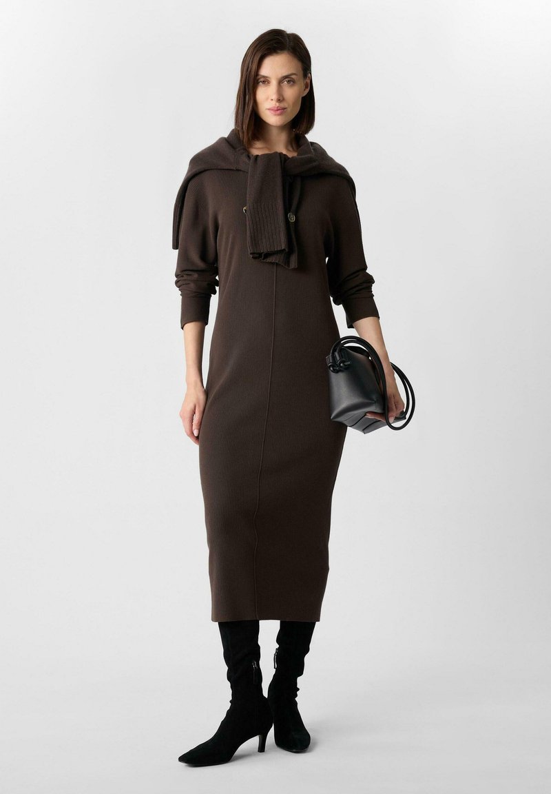 Braunes, geripptes Midikleid mit hohem Ausschnitt und Knopfdetails, kombiniert mit einer schwarzen Handtasche und kniehohen schwarzen Stiefeletten mit Absatz.