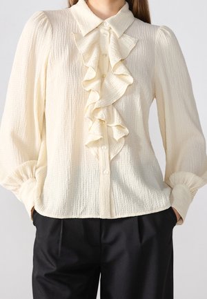 Kermainen tekstuuripusero, jossa röyhelöinen etuosa, puhditut pitkät hihat, napilliset kalvosimet ja kaulus, yhdistetty mustiin housuihin.