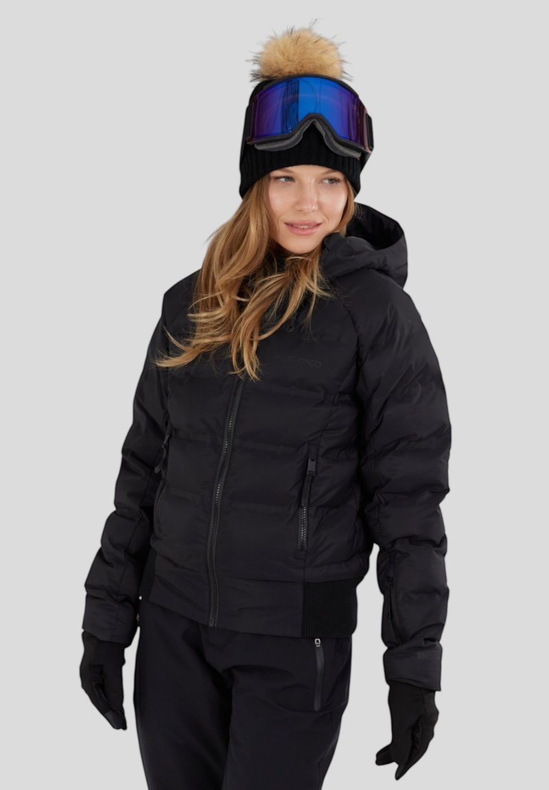 Svart puffjacka med huva, ribbade ärmslut och dragkedjor; bärs med en svart mössa som har en beige pom-pom och blåtonade skyddsglasögon.